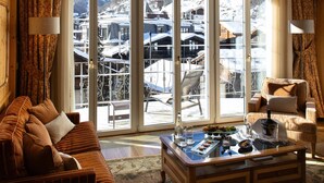 A cozy living room with a large window, a chandelier, and a view of a snowy mountain village. - Mont Cervin Palace (Zermatt)