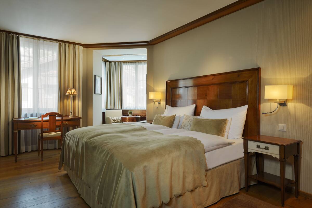 A hotel room with a large bed, a desk, and a chair. There are two lamps on the bedside table and a window with curtains.