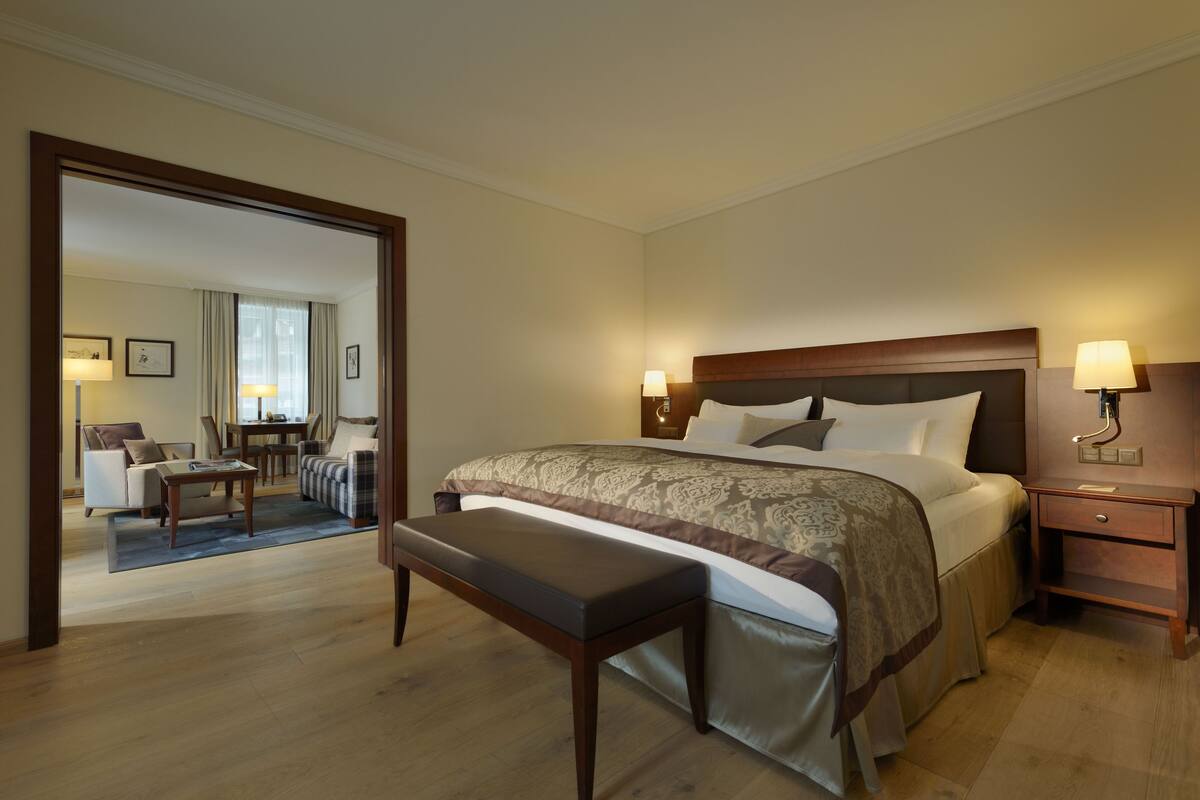 A hotel room with a large bed, bedside tables, a bench, and a view into the living area.