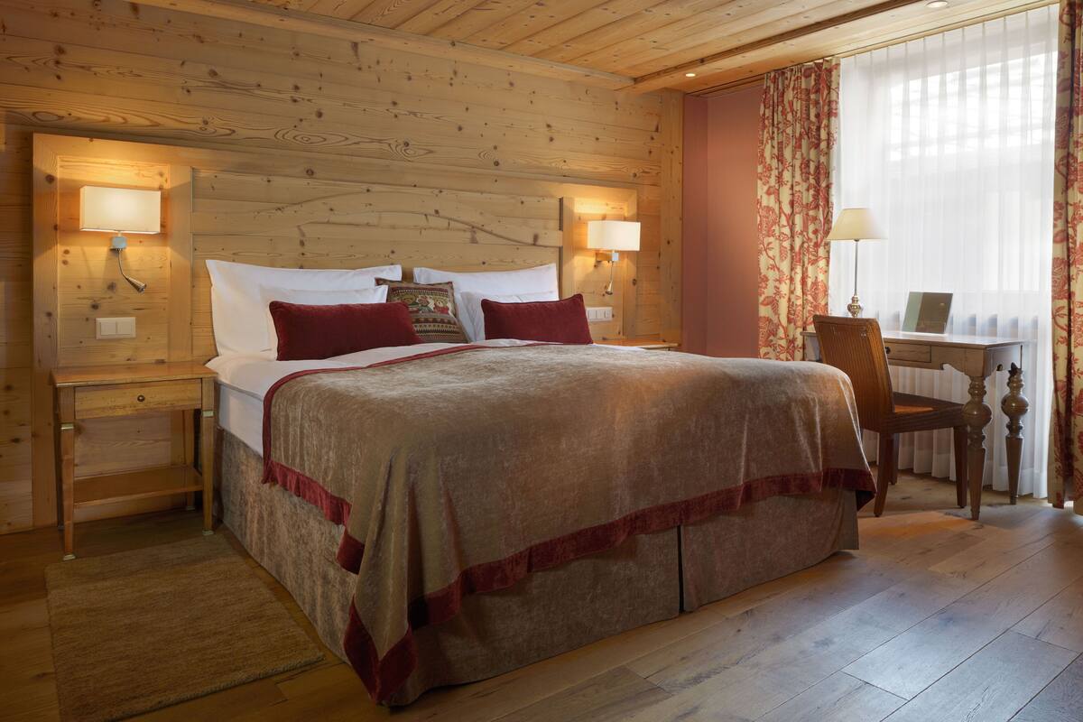 A wooden-paneled bedroom with a bed, two bedside lamps, a desk, and a chair.