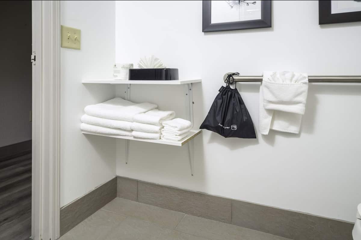 A bathroom with a towel rack, a shelf with folded towels, a black bag, and a framed picture on the wall.