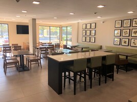 Un café moderno con varios tipos de asientos, como mesas y cabinas, grandes ventanales que dejan entrar luz natural y una pared decorada con impresiones botánicas enmarcadas.