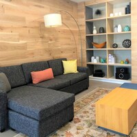 Una sala moderna con un sofá seccional gris, piso de madera, una lámpara de pie, una mesa de centro de madera y un estante con objetos decorativos.