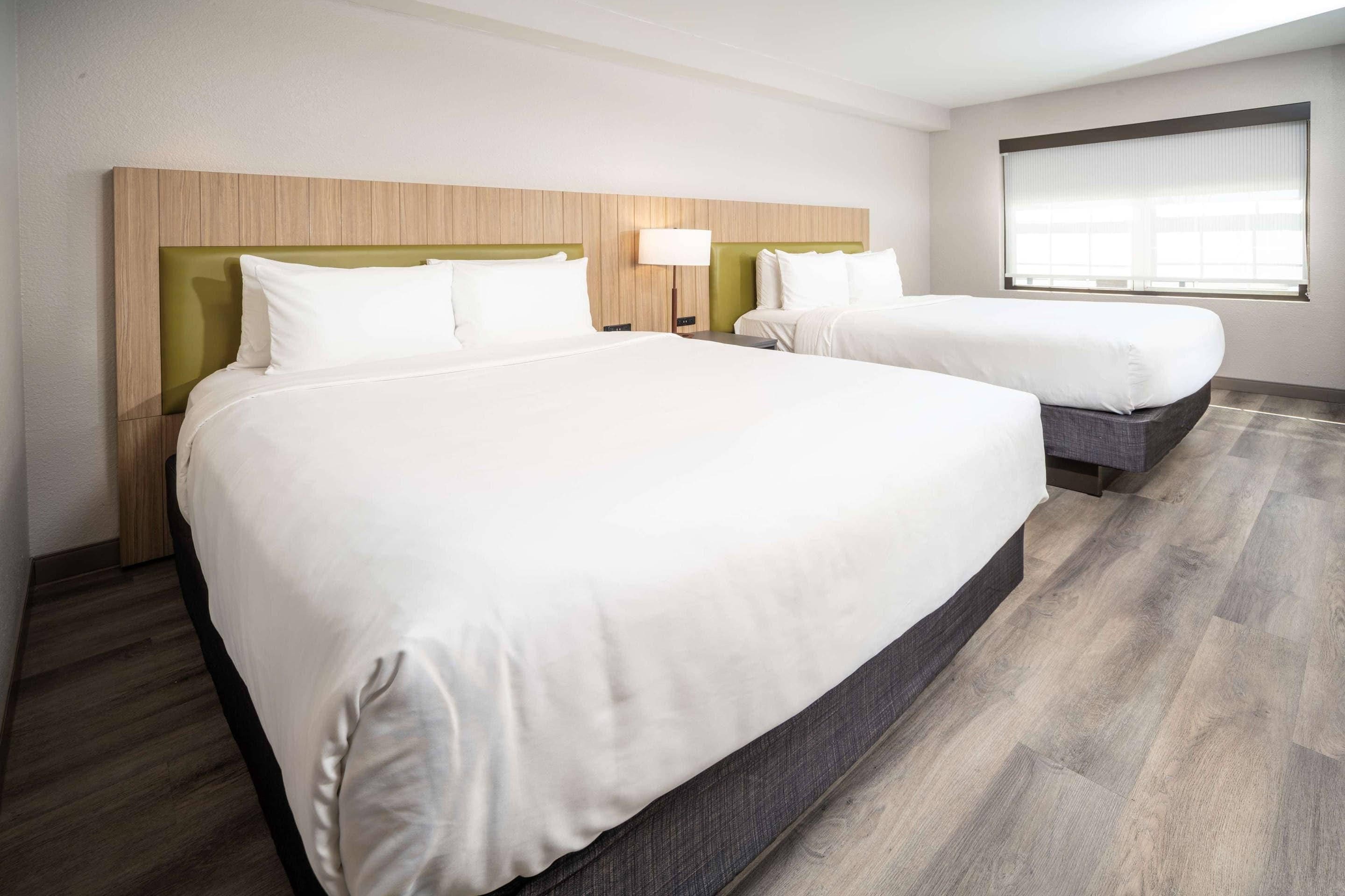 A hotel room with two beds, a wooden headboard, a bedside table with a lamp, and a window with blinds.