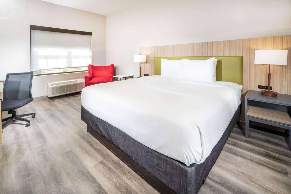 A hotel room with a large bed, a desk, a red chair, and a window with blinds.