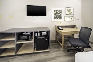 Habitación de hotel con televisor de pantalla plana, escritorio con computadora, silla, microondas y refrigerador pequeño.