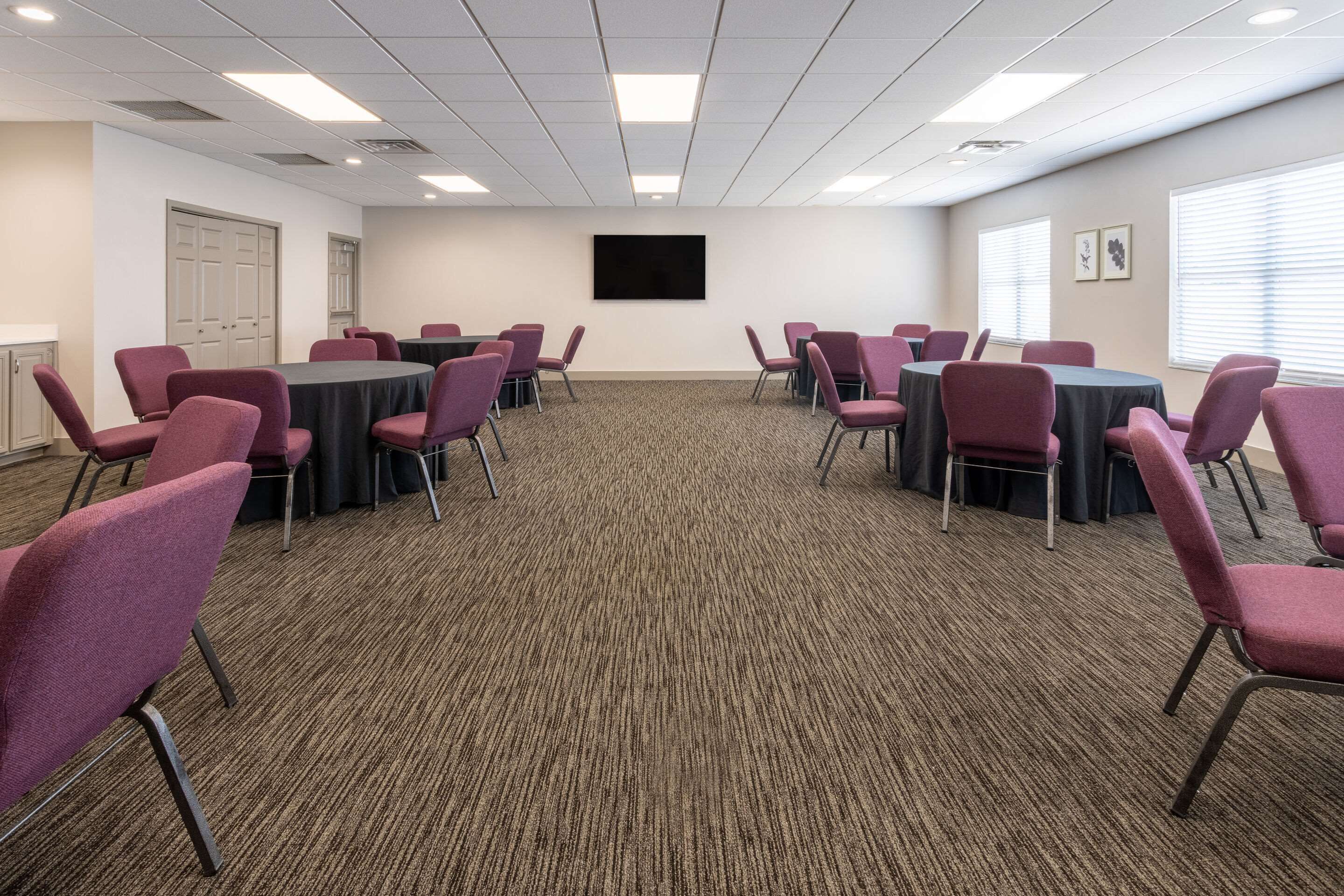 Une salle de conférence avec des tables rondes et des chaises violettes, un sol recouvert de moquette, des murs blancs et un téléviseur à écran plat fixé au mur.