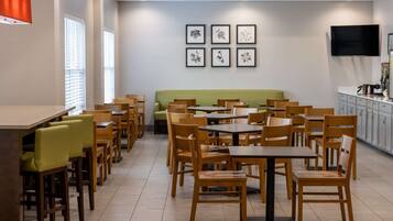 A modern dining area with wooden tables and chairs, a green sofa, and a flat-screen TV mounted on the wall.