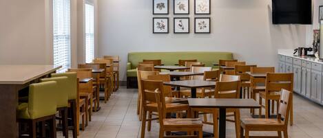 A modern dining area with wooden tables and chairs, a green sofa, and a flat-screen TV mounted on the wall.