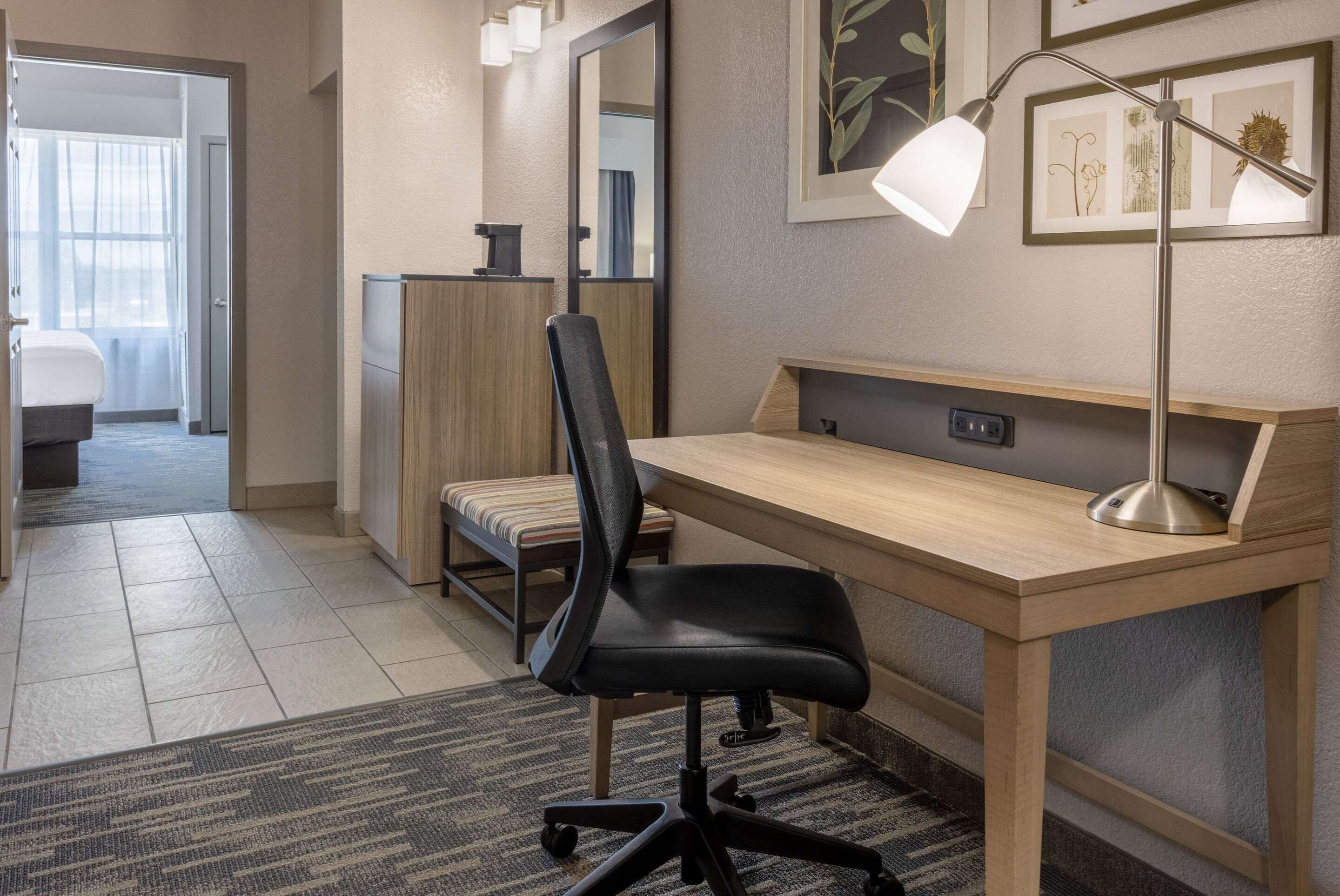 A modern office space with a wooden desk, black office chair, and a standing lamp. There is a framed artwork on the wall and a small shelf with a decorative item.