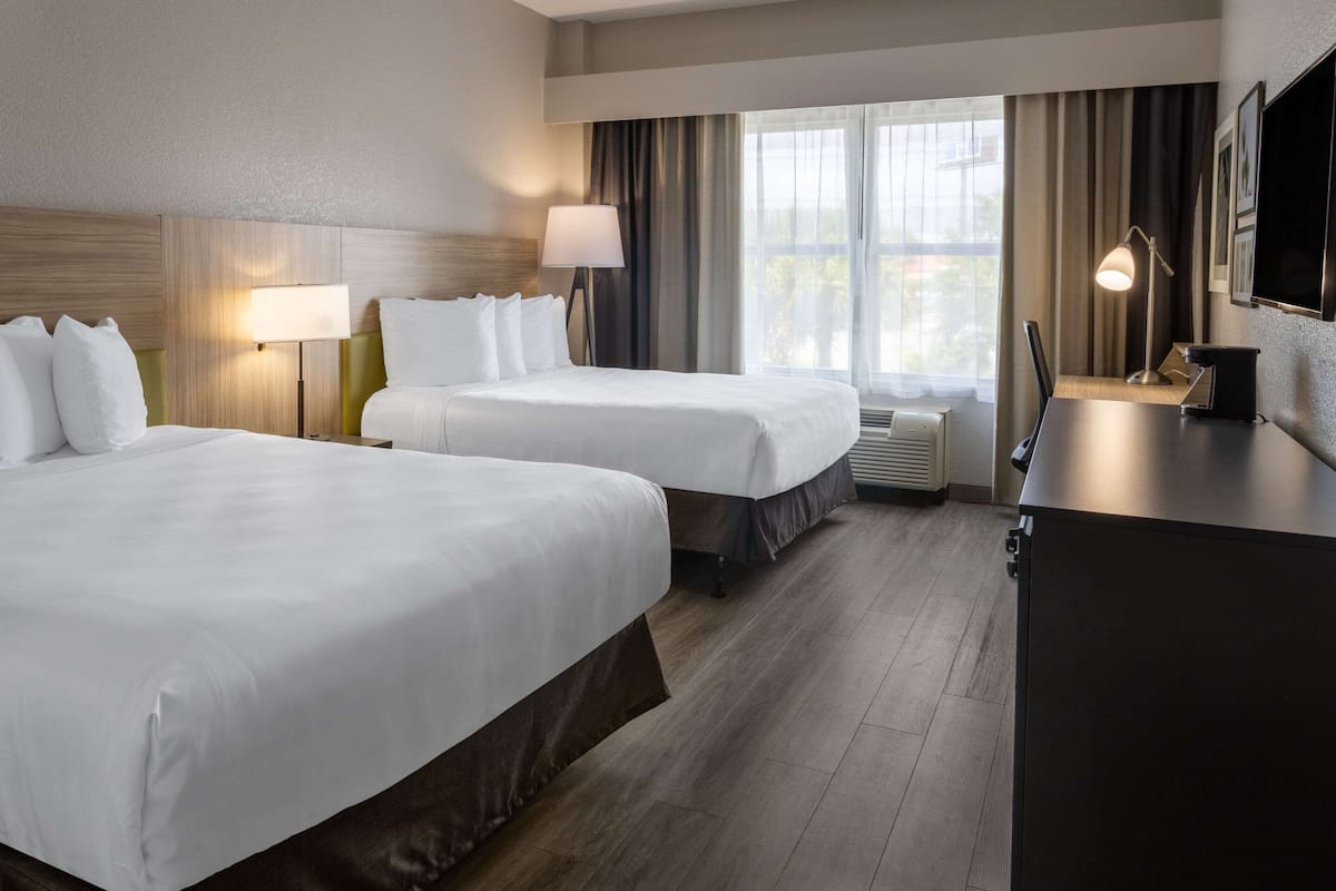A hotel room with two beds, a desk, a lamp, and a large window with curtains.