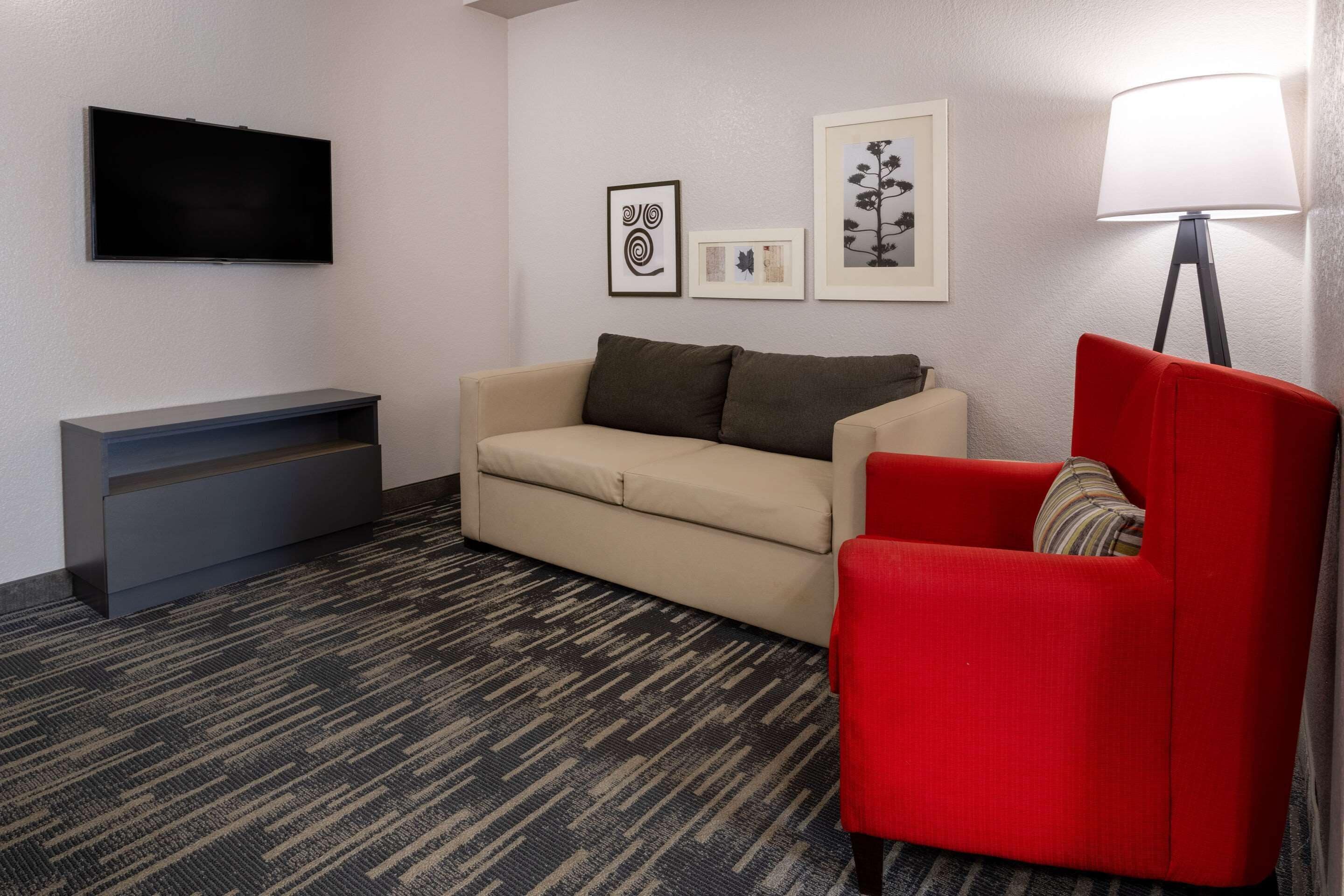 Una sala de estar con un sofá color beige, un sillón rojo, un televisor de pantalla plana, una pequeña consola y cuadros en la pared.