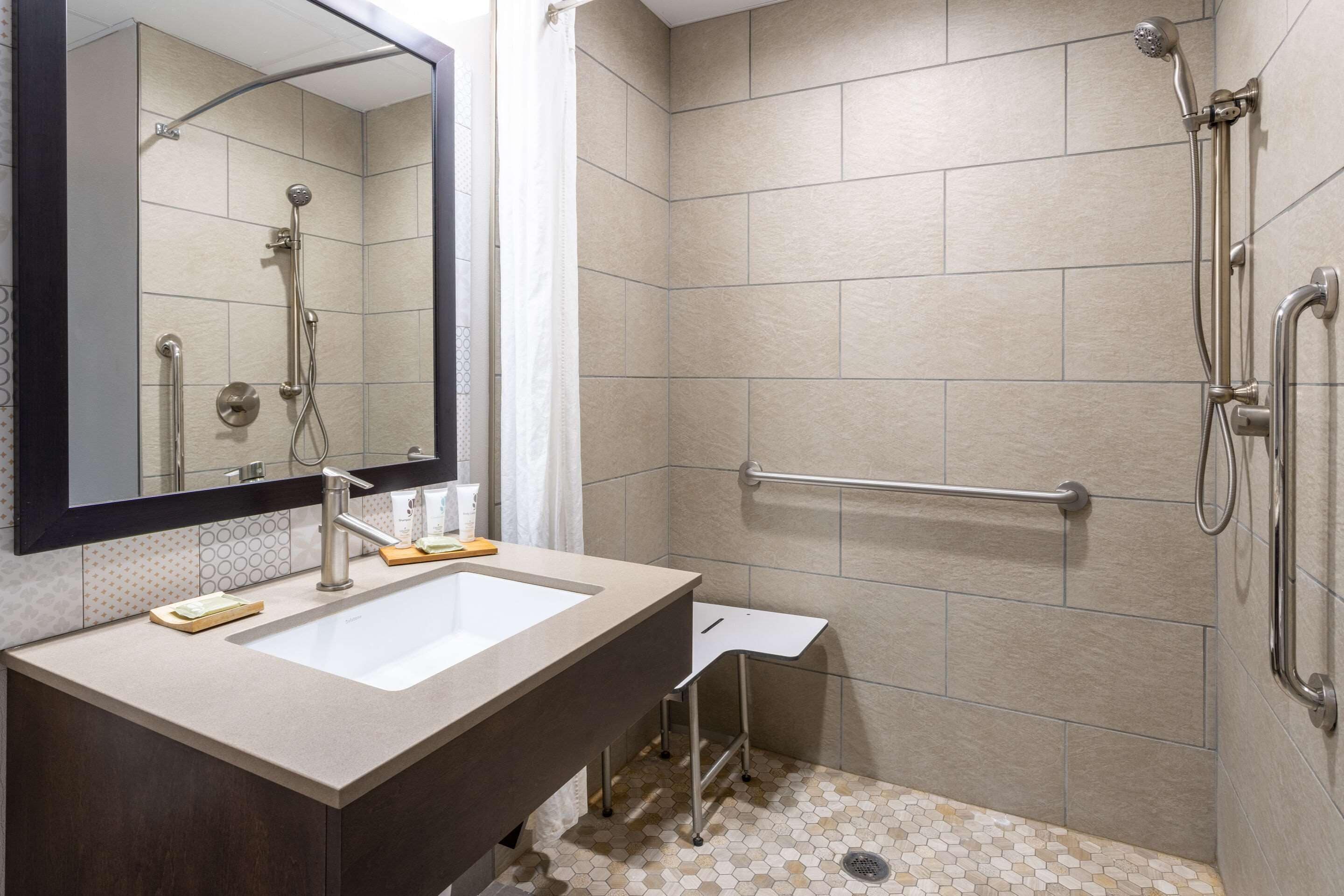 Un baño moderno con un espejo grande, un lavamanos, una ducha con cabezal de ducha manual y una barra de apoyo.