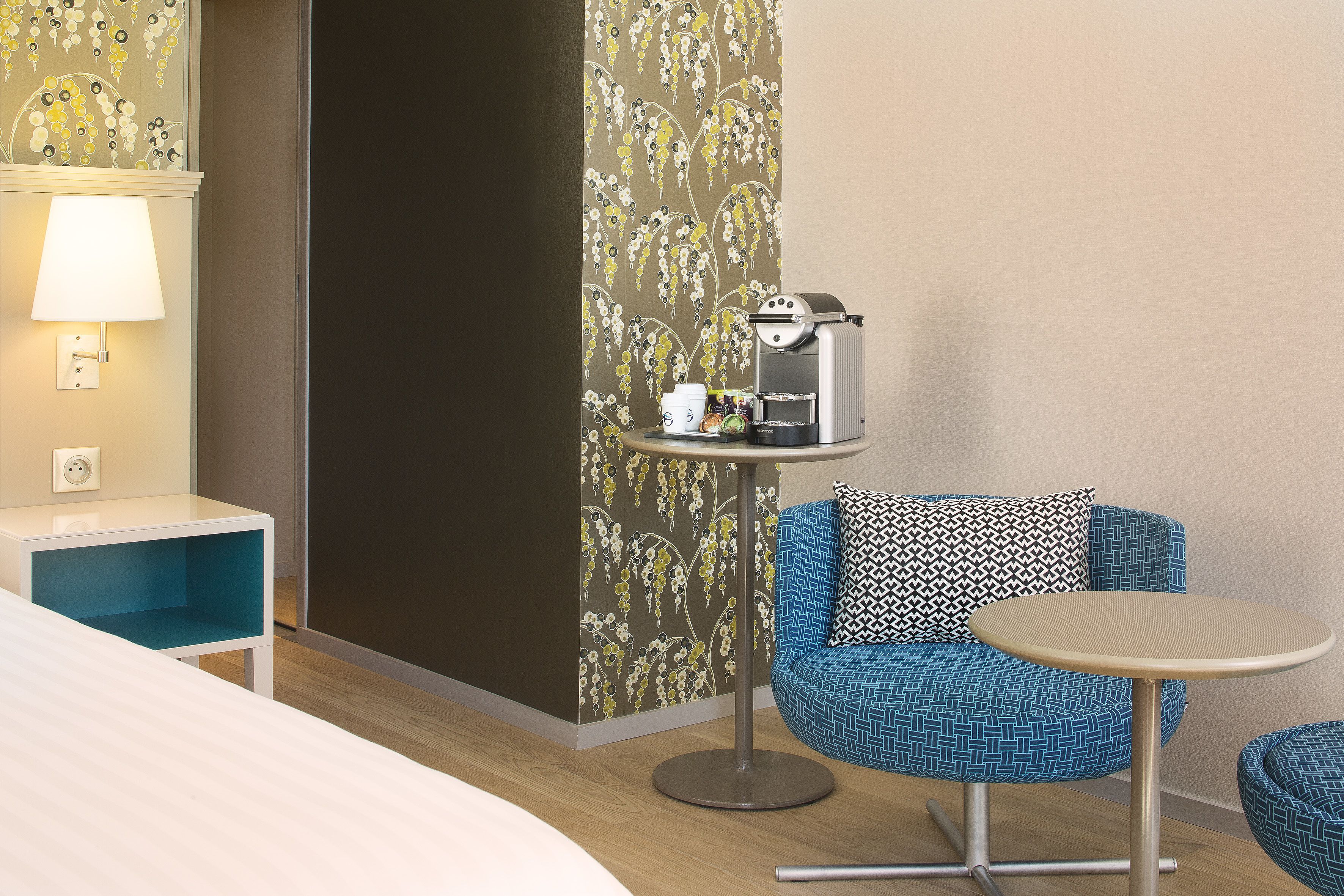 A hotel room with a bed, a small table with a coffee maker, a blue chair, and a wall with a floral pattern.