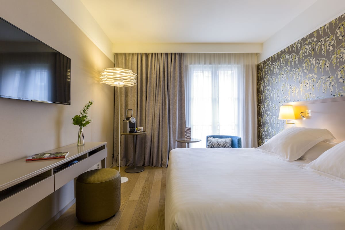 A hotel room with a bed, a TV mounted on the wall, a desk with a lamp, and a window with curtains.