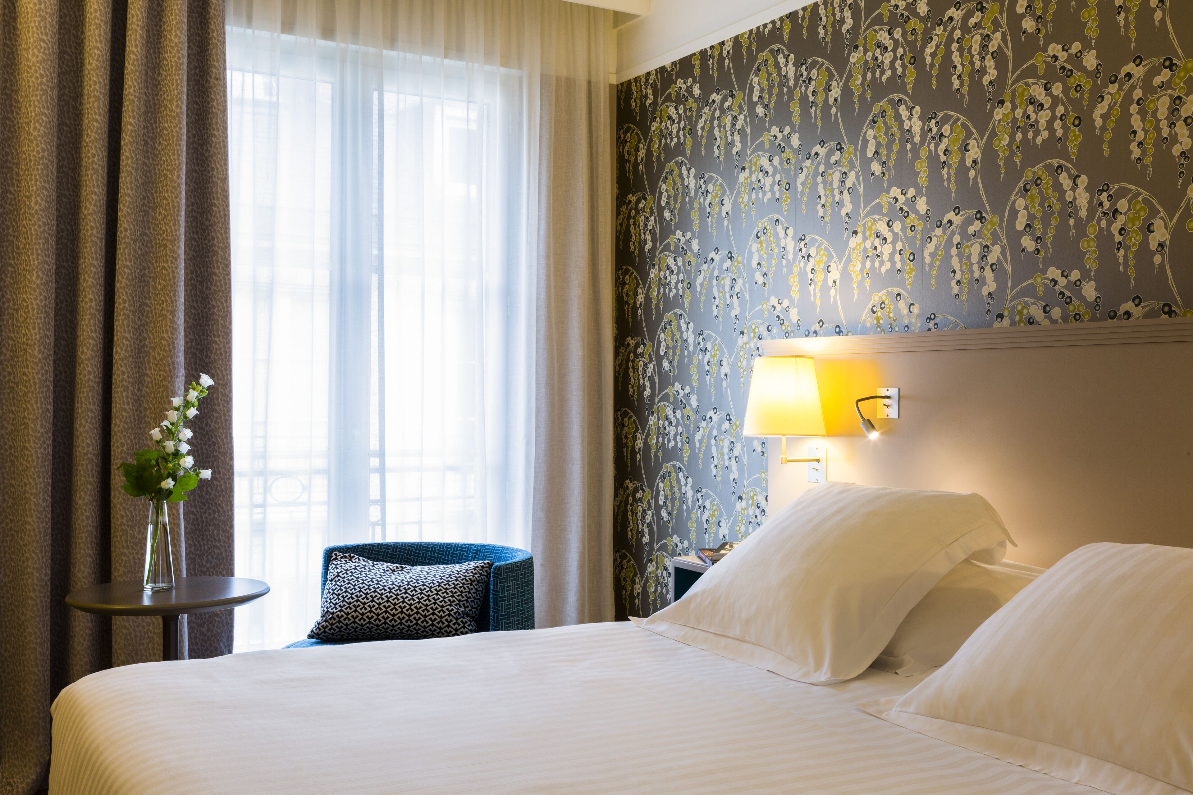 A hotel room with a large bed, a small round table with a vase of flowers, a chair with a patterned cushion, and a wall with a floral wallpaper.