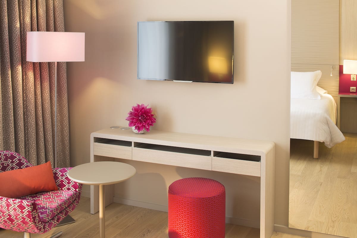A modern hotel room with a flat-screen TV mounted on the wall, a small desk with drawers, a round table, a red cylindrical ottoman, a patterned chair with an orange cushion, and a bed with white bedding.