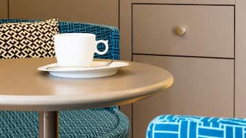 Um quarto com uma mesa redonda, uma chávena branca num pires, uma cadeira azul com padrão e um minibar, além de uma parede estampada.