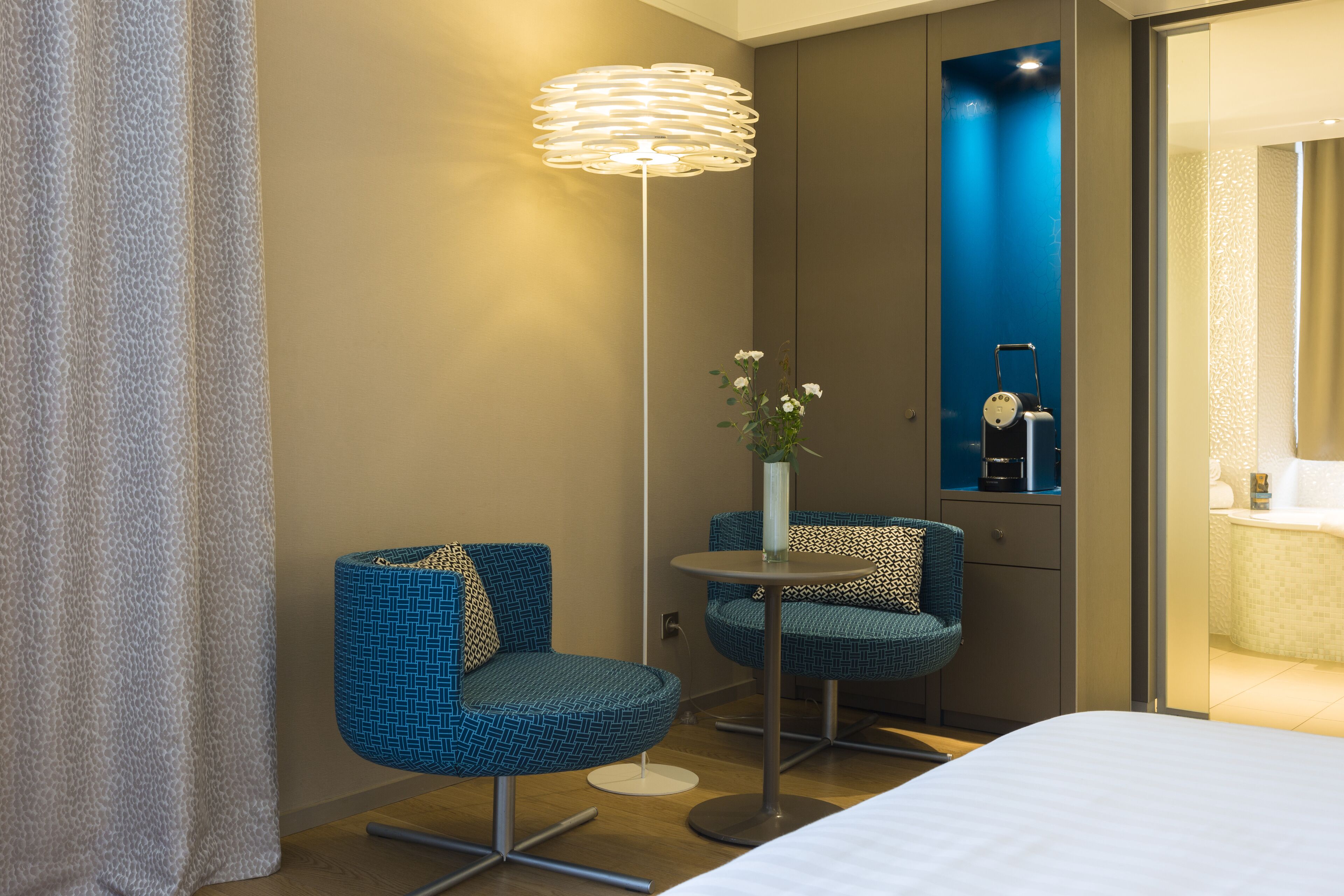 Et hotelværelse med en seng, to blå stole, et lille bord med en blomstervase og en moderne gulvlampe.