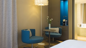 Et hotelværelse med en seng, to blå stole, et lille bord med en blomstervase og en moderne gulvlampe.