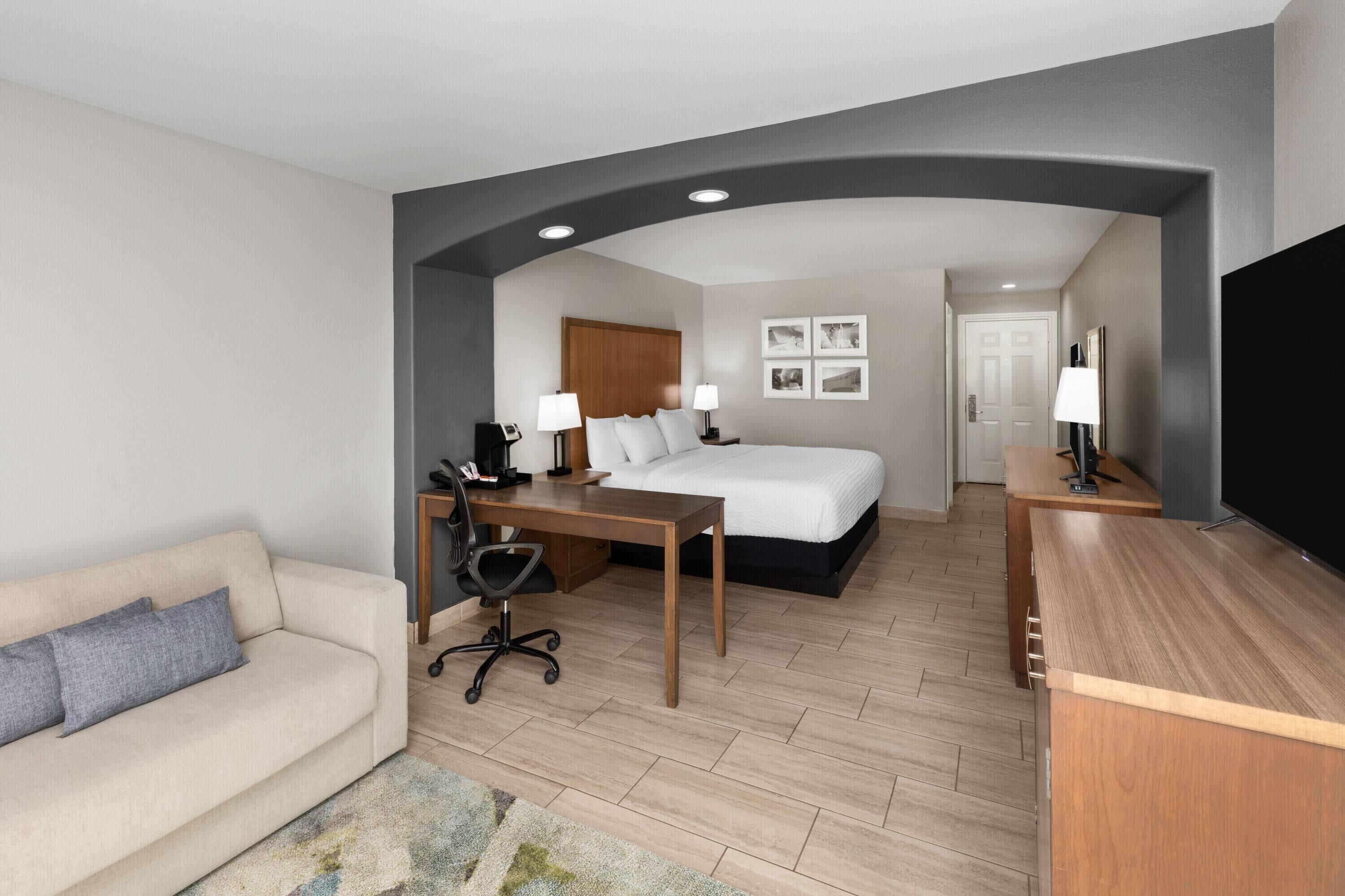 A hotel room with a bed, a desk, a sofa, a TV, and a door leading to another room.