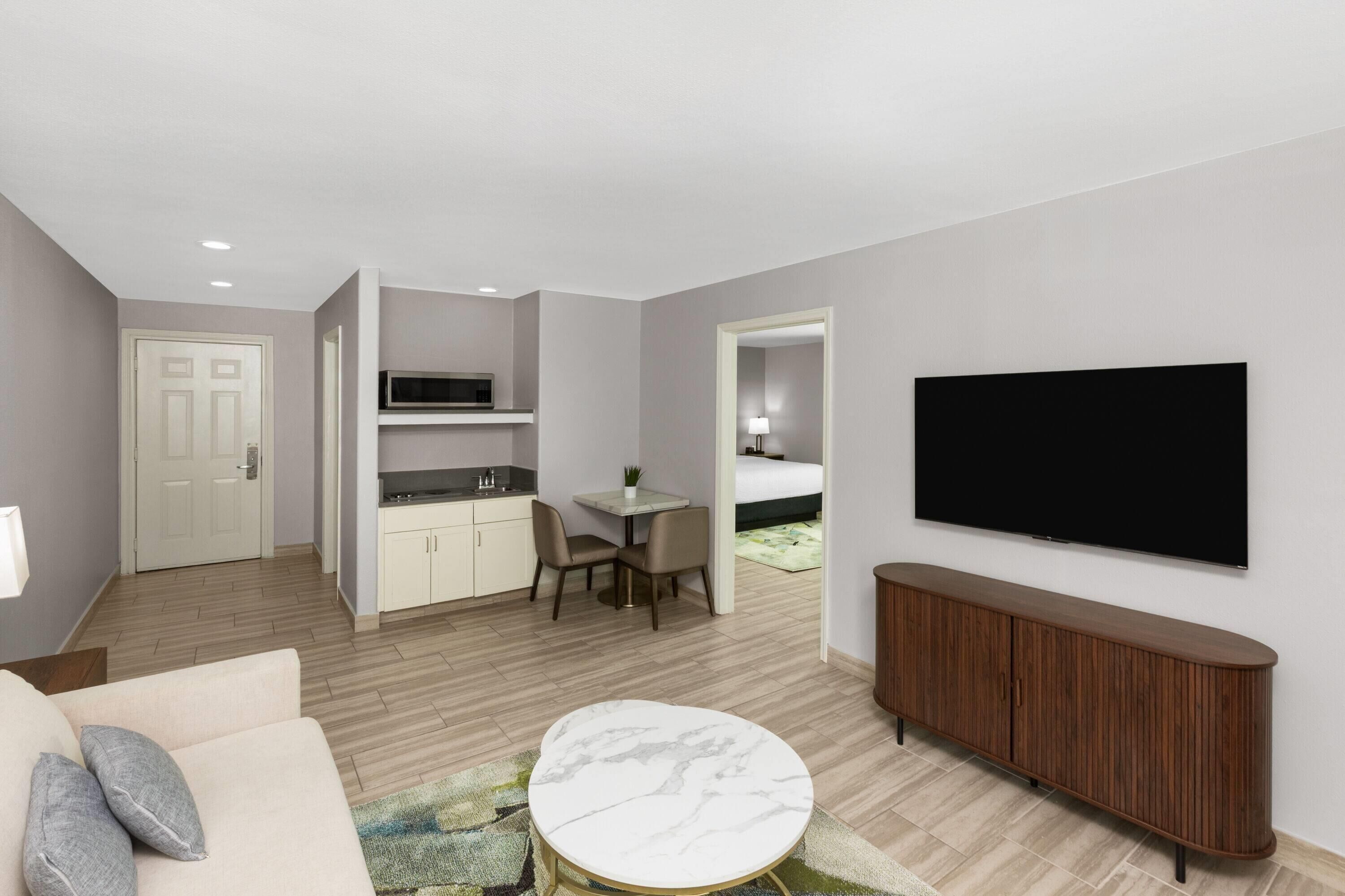 A modern living room with a sofa, a round coffee table, a TV, a wooden cabinet, and a kitchen area with a microwave.