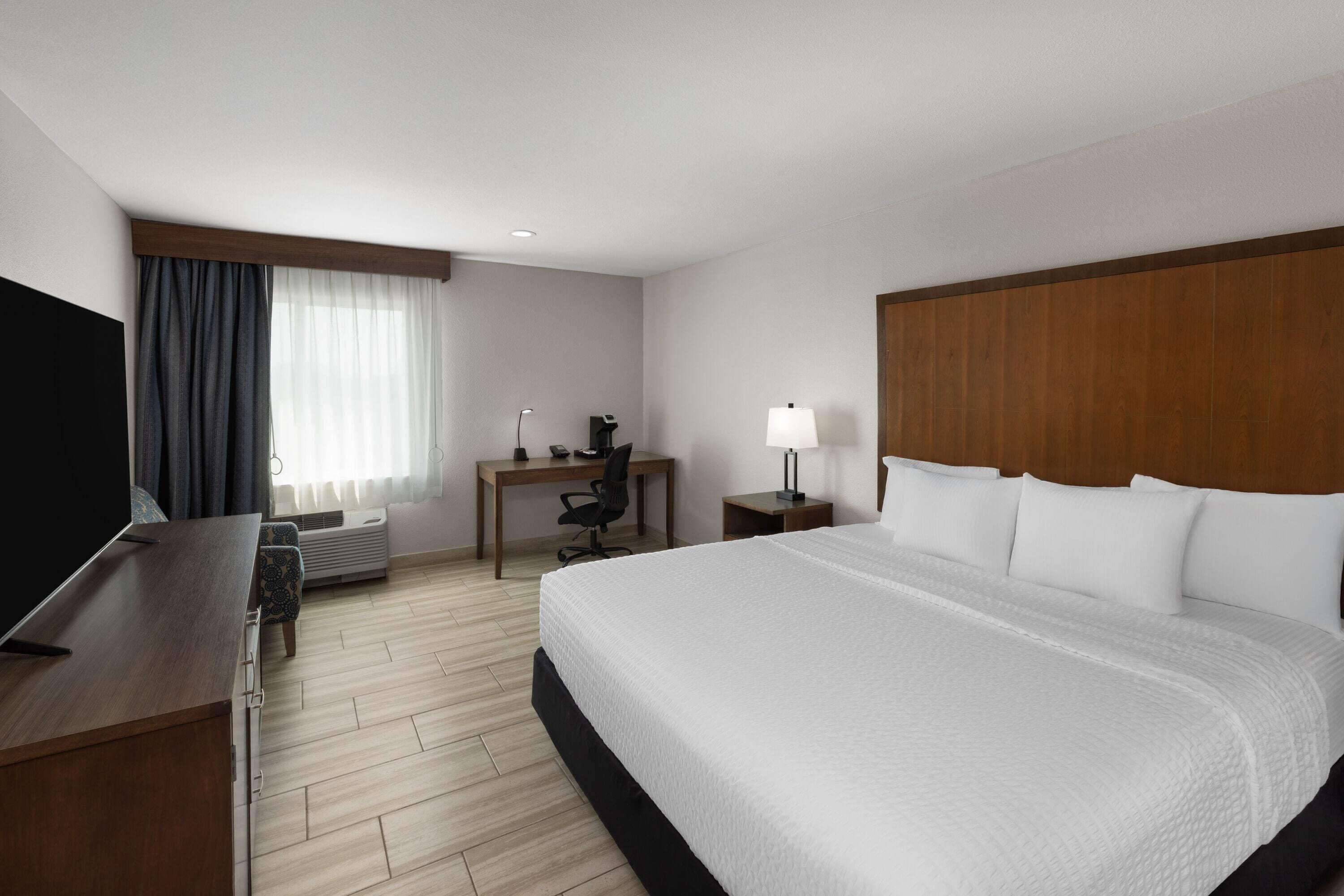 A hotel room with a large bed, a desk, a chair, a TV, and a lamp.