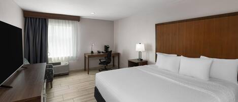 A hotel room with a large bed, a desk, a chair, a TV, and a lamp.