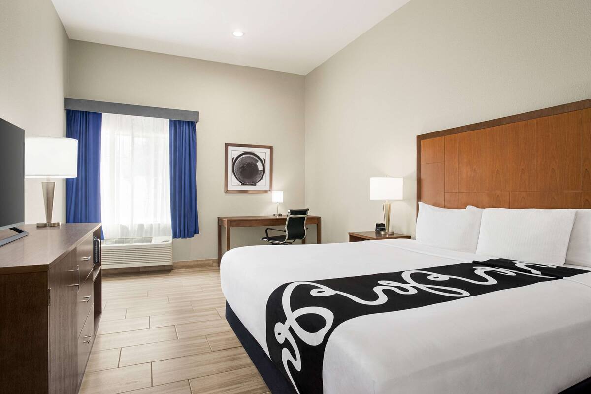 Ein Hotelzimmer mit einem großen Bett, einem Schreibtisch, einem Stuhl, einem Fernseher und einem Fenster mit Vorhängen.