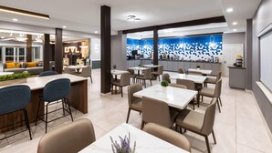 A modern hotel lobby with a bar area, dining tables, and a kitchen in the background. - La Quinta Inn & Suites by Wyndham Brownsville North (Brownsville)
