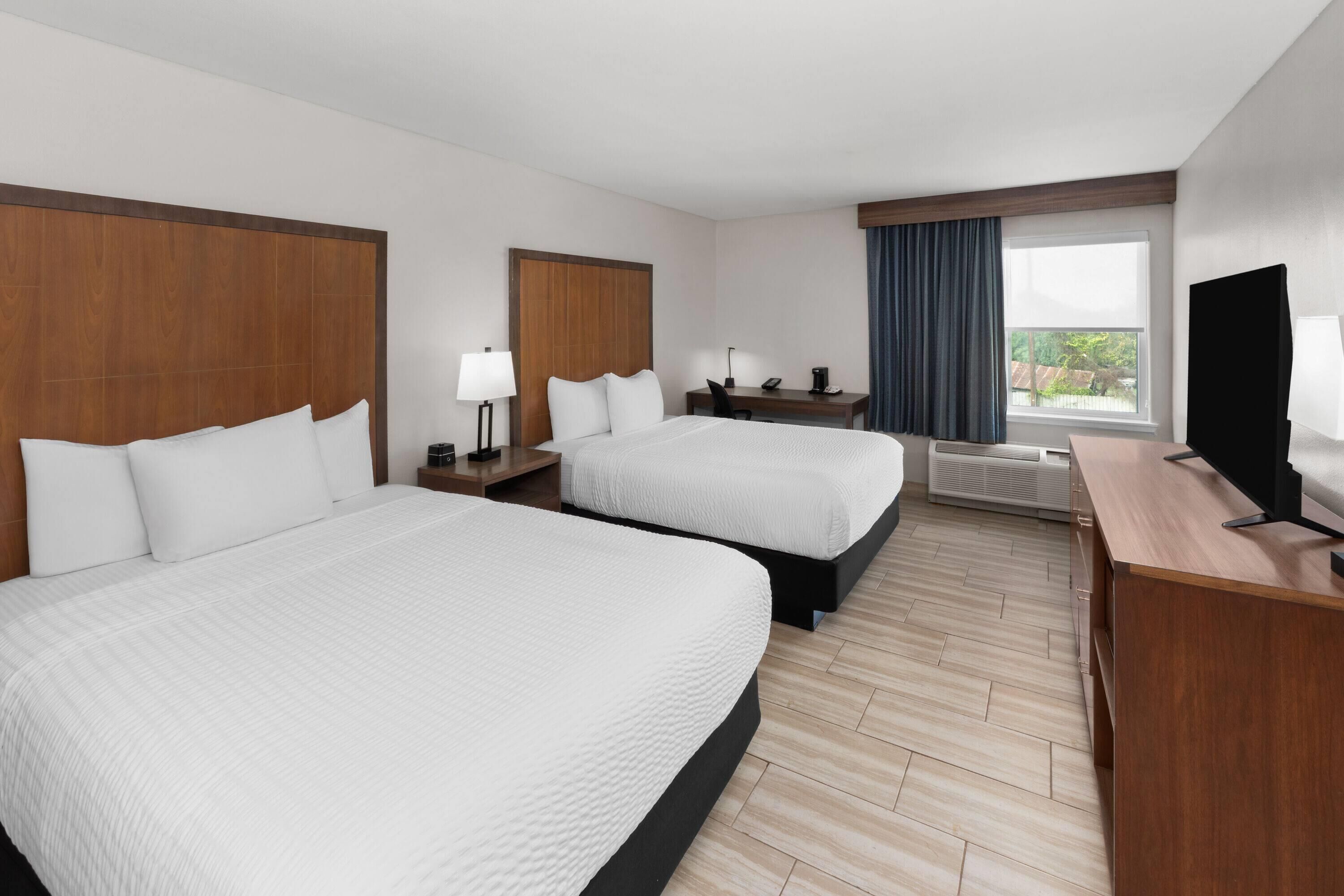 A hotel room with two beds, a desk, a TV, and a window with curtains.