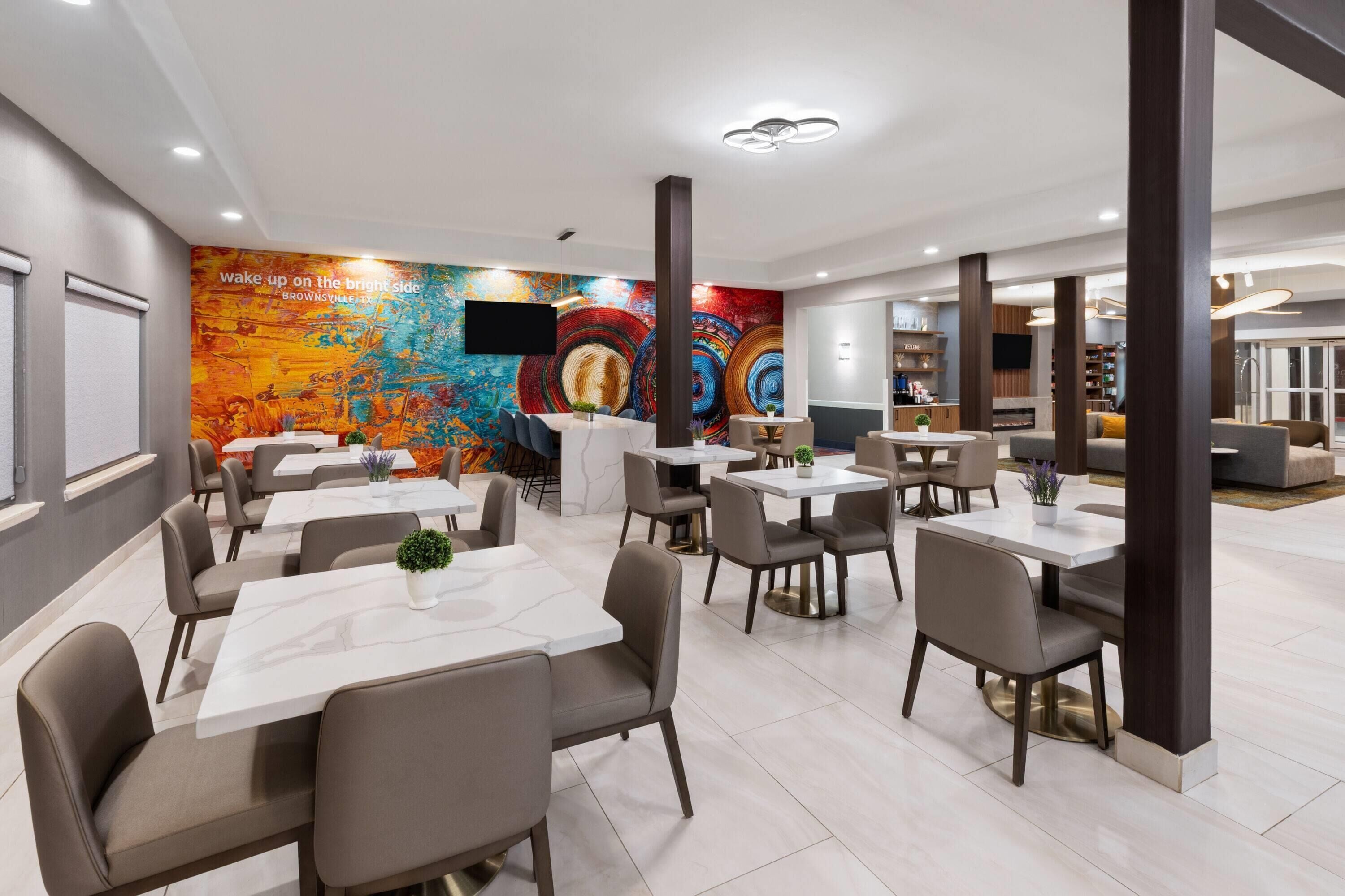 A modern dining area with tables and chairs, a colorful mural, and a flat-screen TV mounted on the wall.