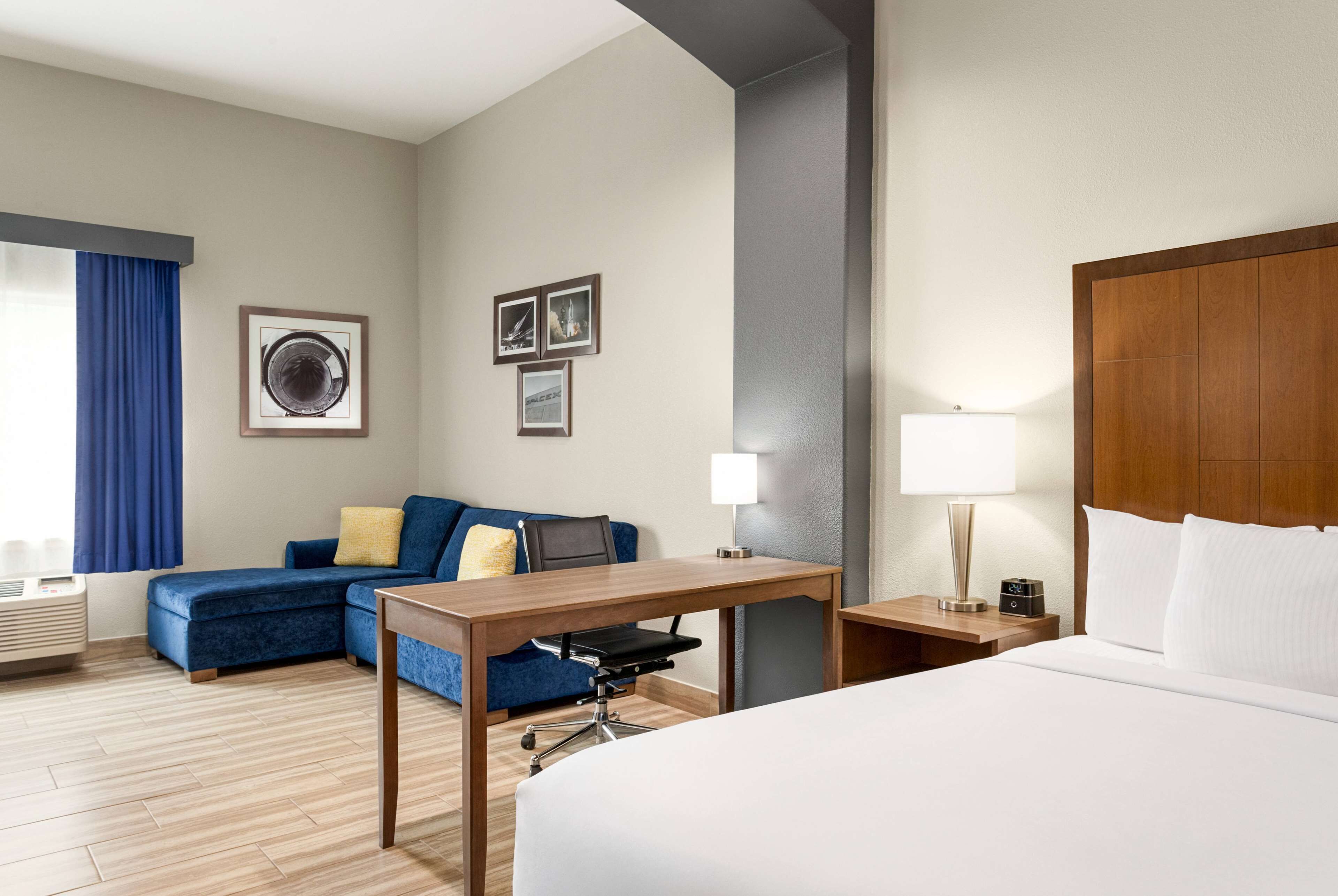 A hotel room with a bed, a desk, a chair, a sofa, a lamp, and a picture frame on the wall.