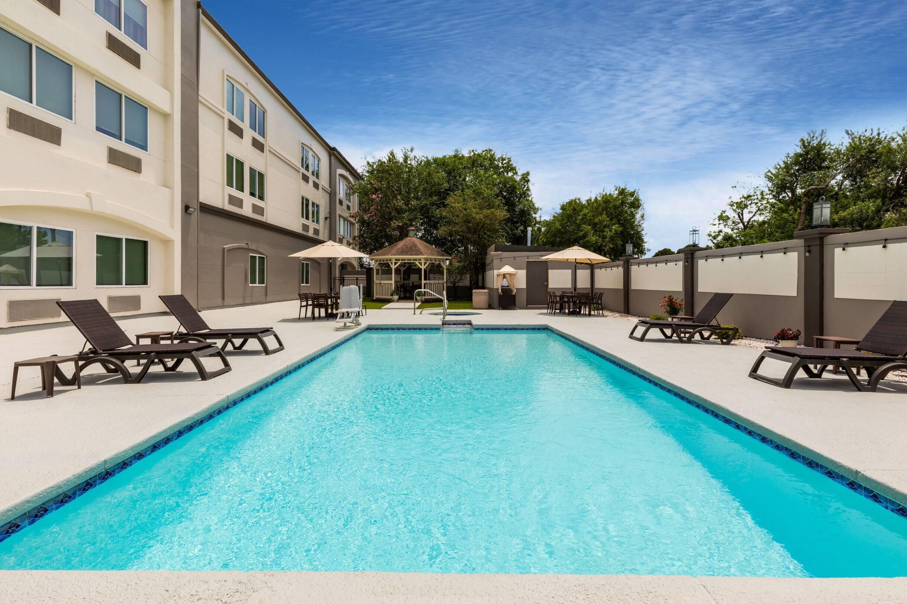 Una piscina rectangular con sillas de descanso y sombrillas, rodeada por un edificio con varias ventanas y un área de estar cubierta.