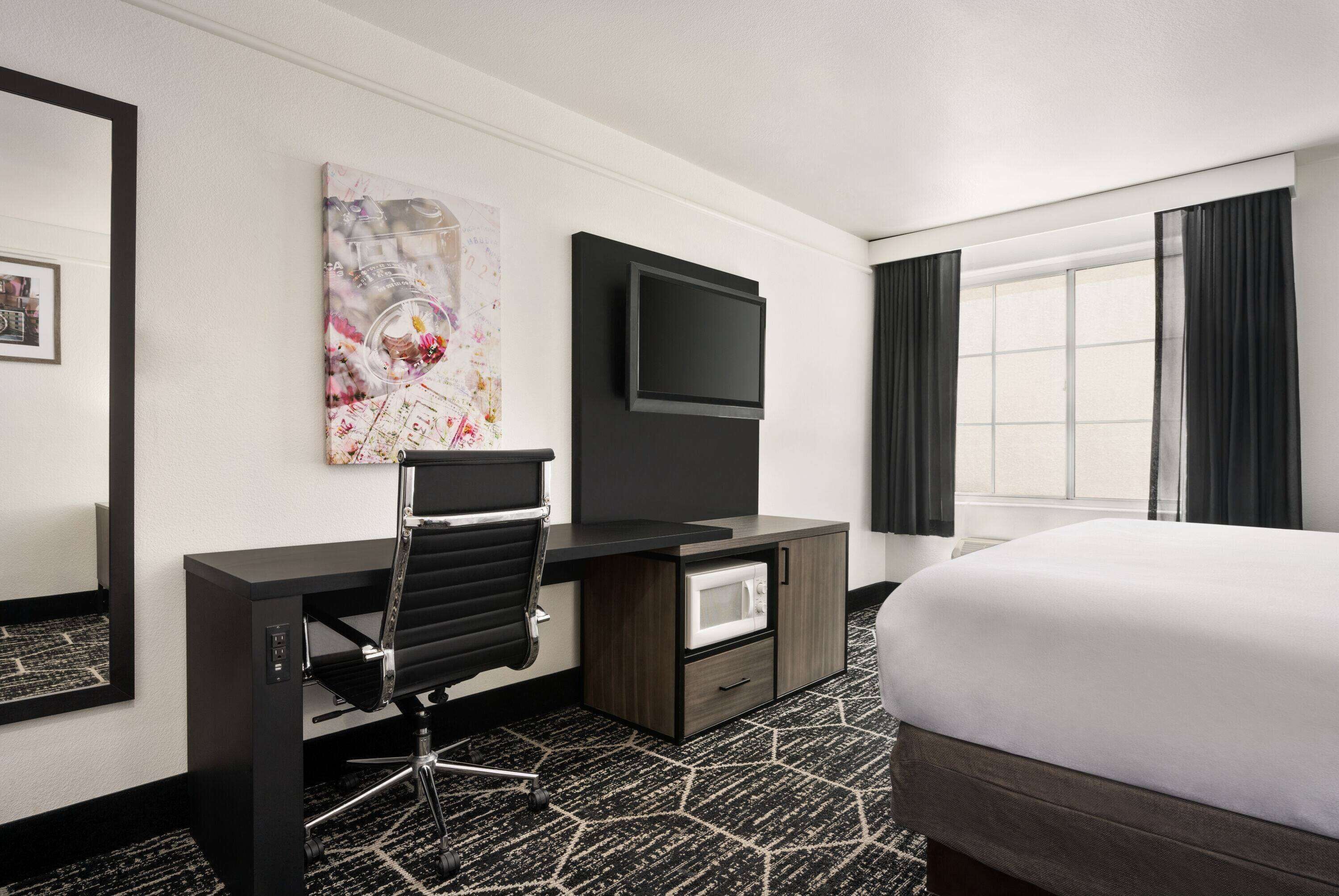A hotel room with a bed, a desk with a chair, a flat-screen TV, a microwave, and a large window with curtains.