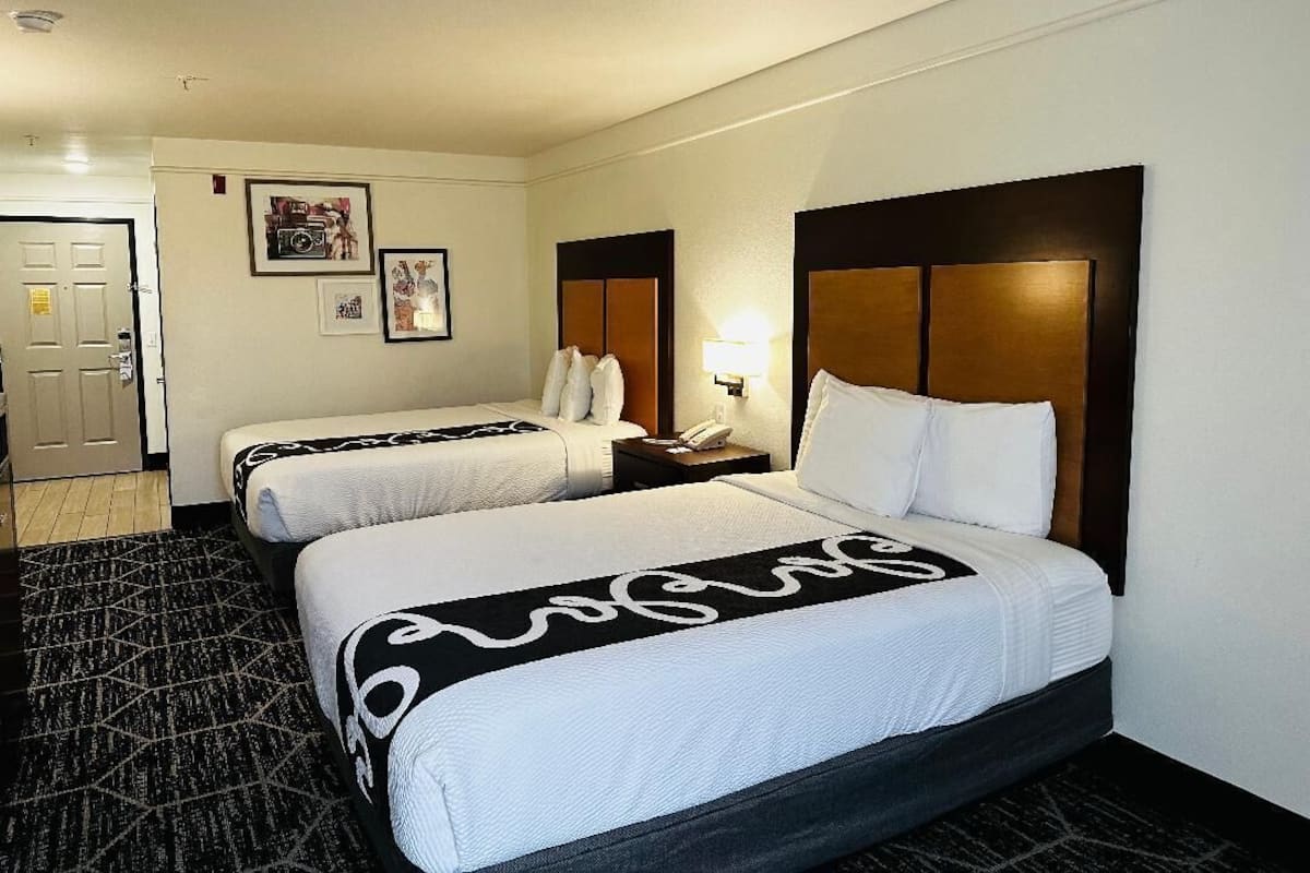 A hotel room with two beds, a nightstand with a lamp, and a door leading to a bathroom.