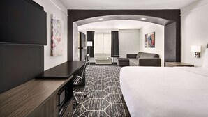 Habitación de hotel con una cama grande, un escritorio, una silla y un sofá. Tiene una ventana con cortinas, un cuadro en la pared y una alfombra con diseño geométrico.