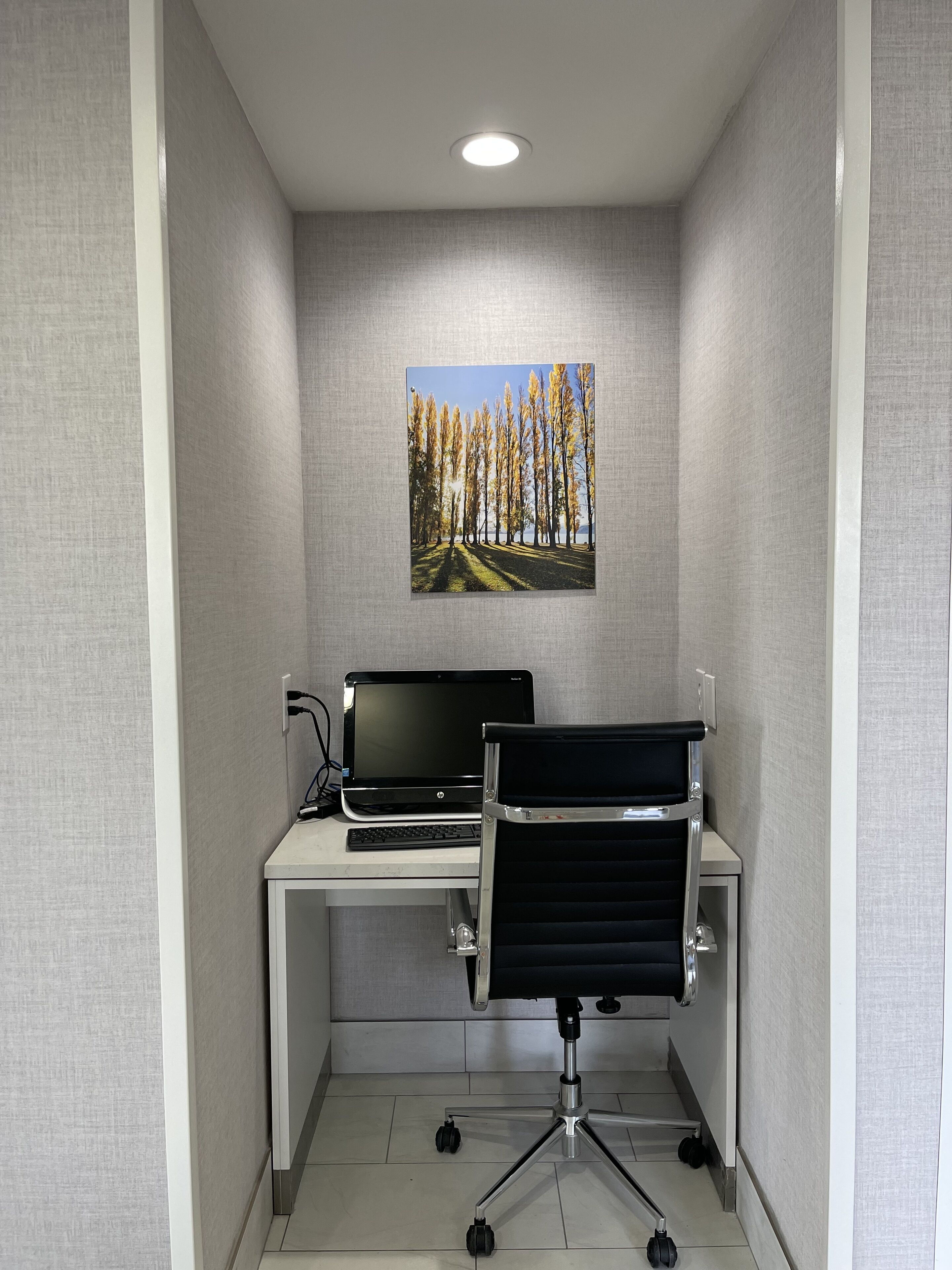 A small office space with a white desk, a black office chair, a laptop, and a framed picture of trees on the wall.