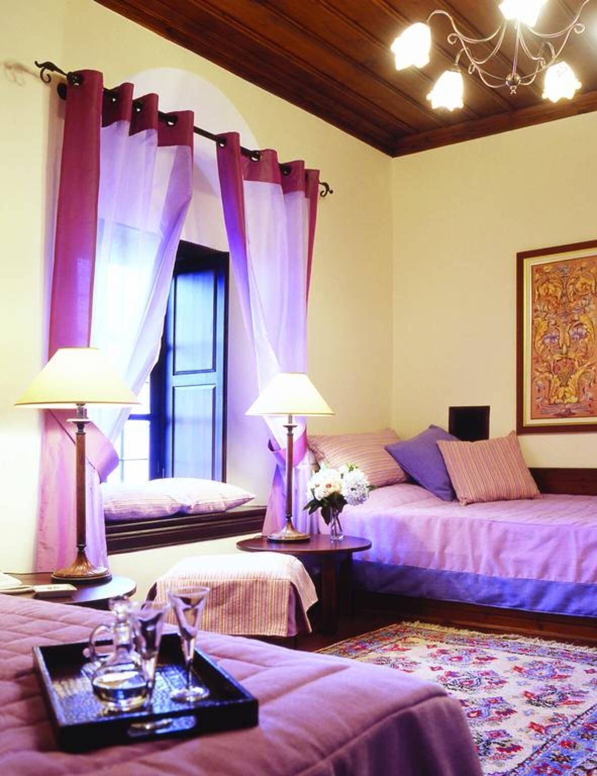 A hotel room with two beds, a chandelier, a painting, and a tray with glasses.