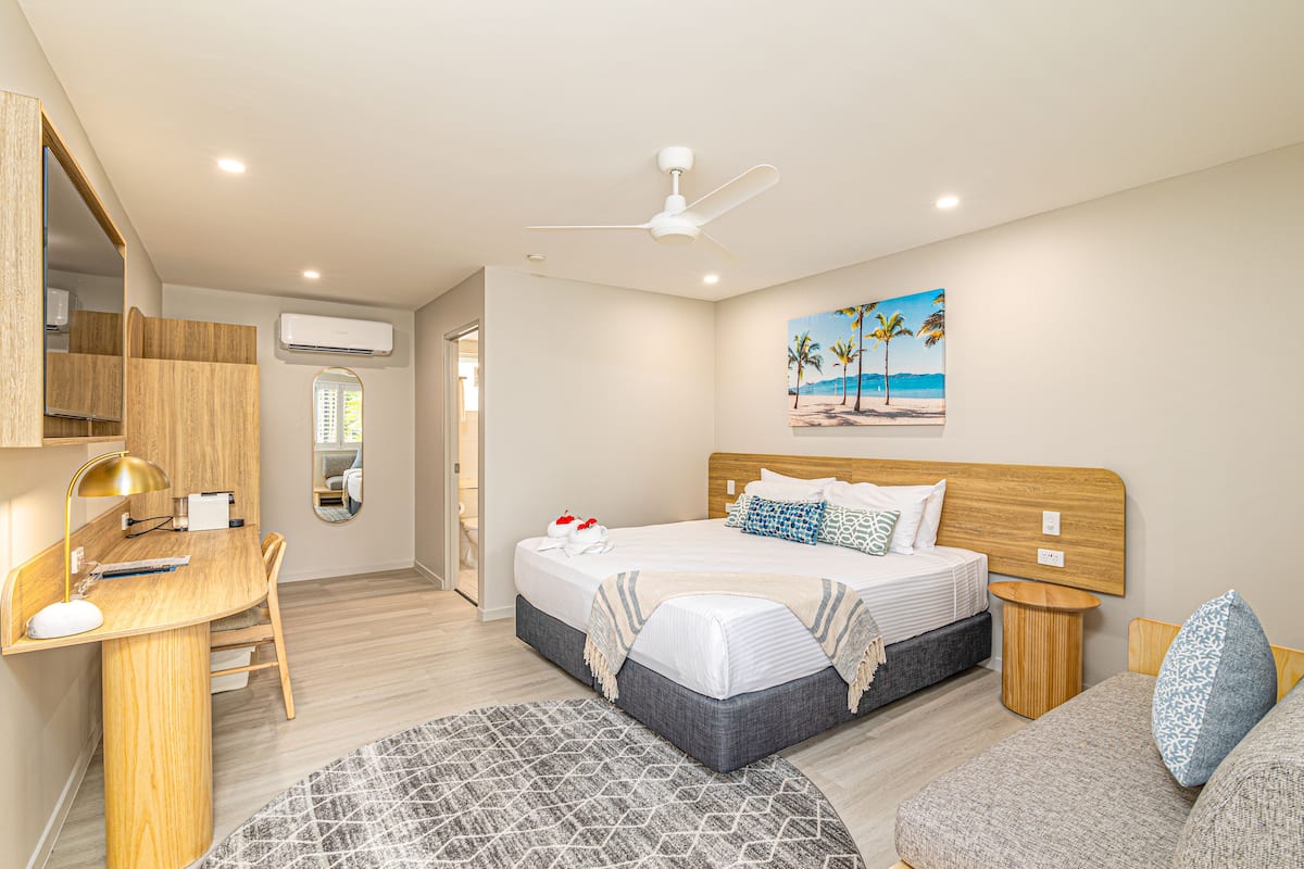 A modern bedroom with a bed, desk, and chair, a ceiling fan, and a beach-themed painting on the wall.