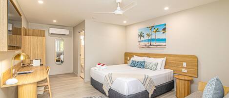A modern bedroom with a bed, desk, and chair, a ceiling fan, and a beach-themed painting on the wall.