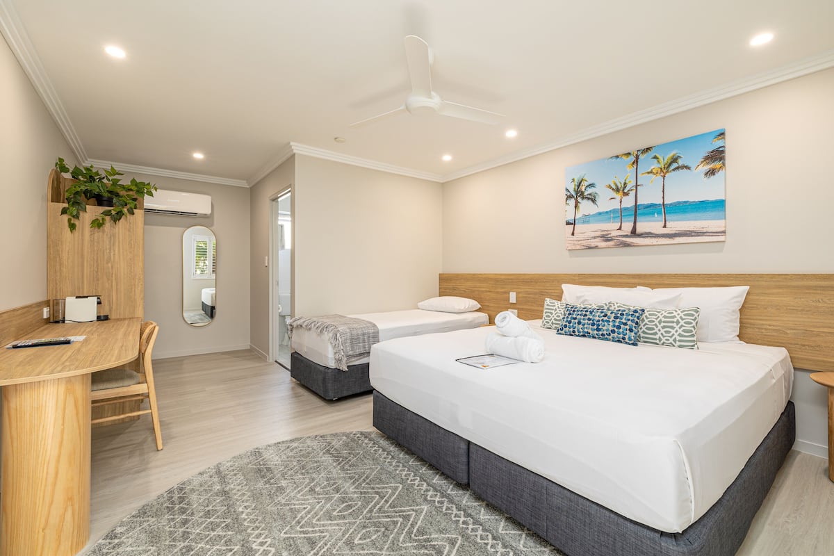 A modern hotel room with a large bed, a desk, a chair, a ceiling fan, and a beach-themed painting on the wall.