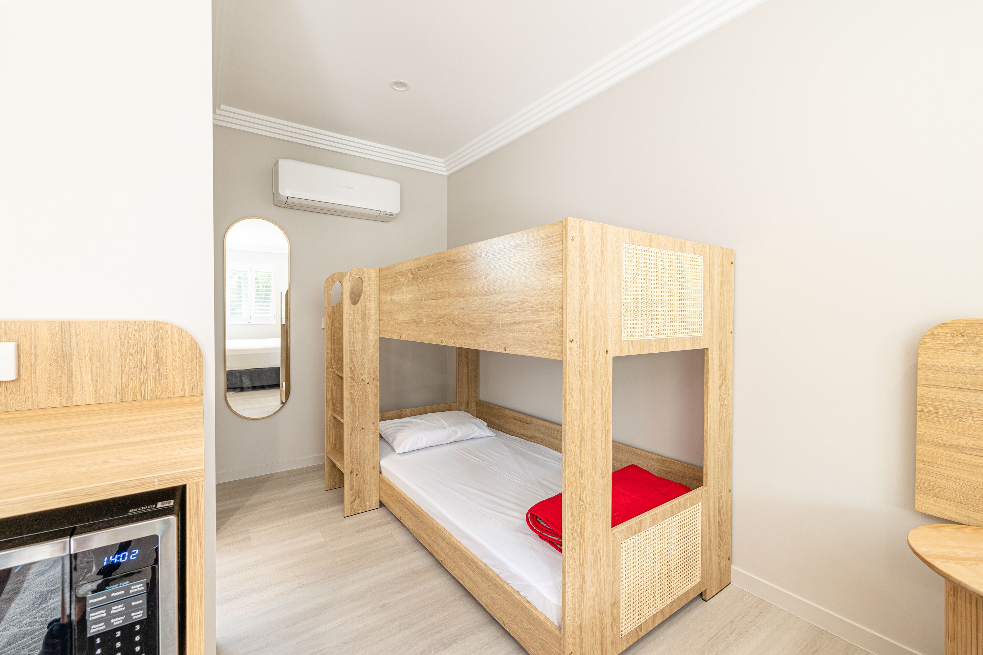 Uma cama beliche com travesseiro vermelho, uma escrivaninha de madeira e um ar condicionado fixado na parede.