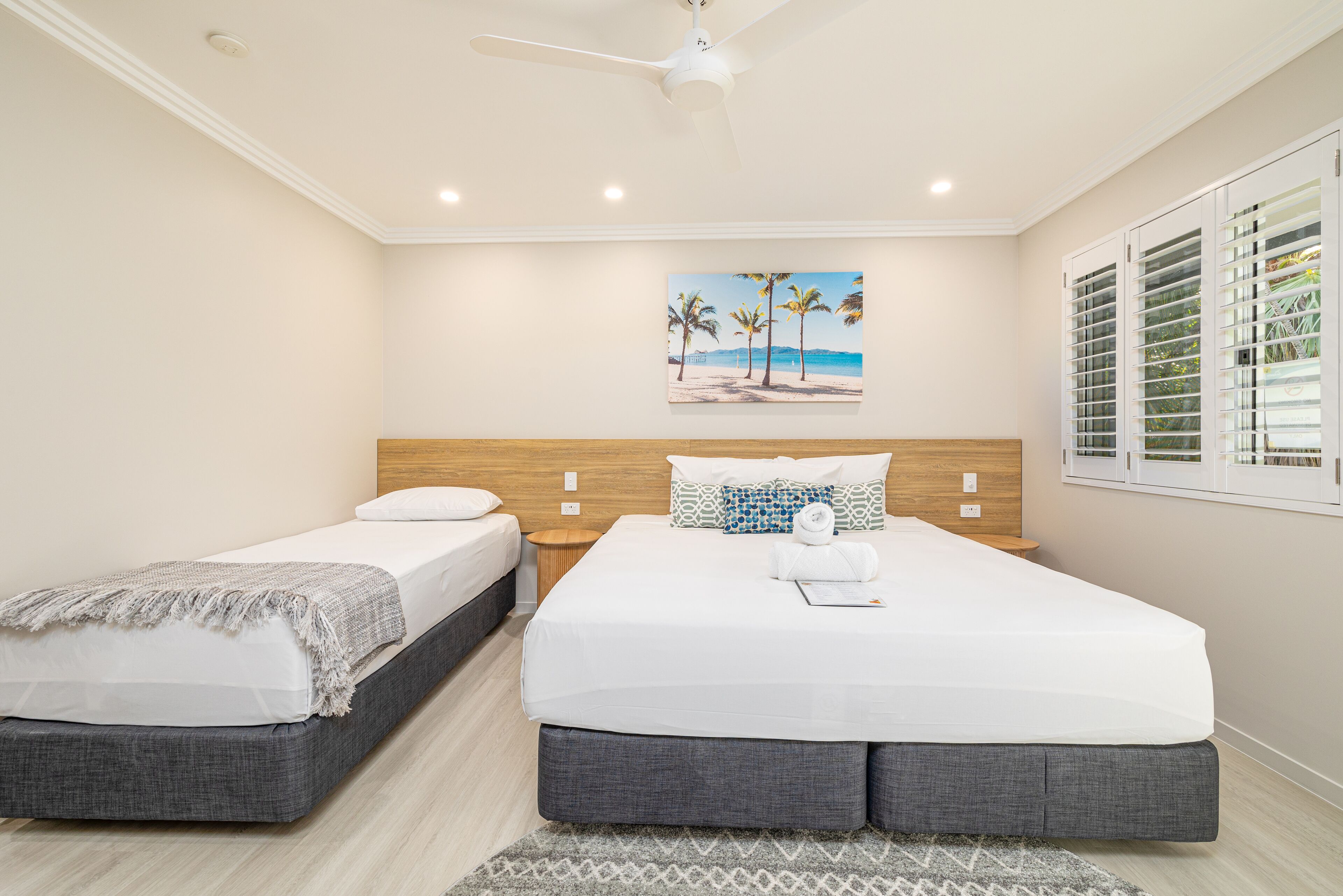 A hotel room with two beds, a ceiling fan, a beach-themed painting, and a window with blinds.