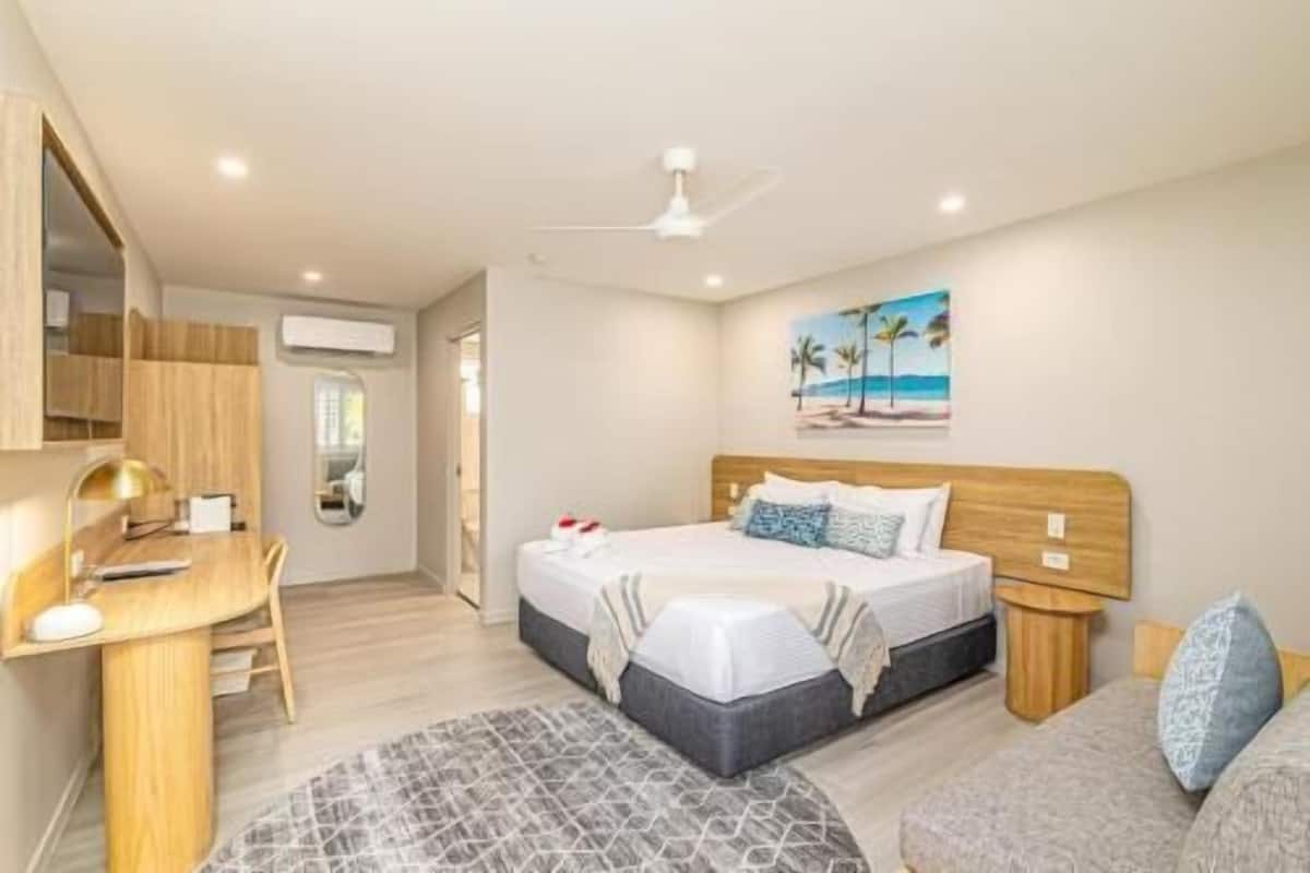 A bedroom with a bed, bedside table, a desk with a chair, a ceiling fan, and a painting on the wall.