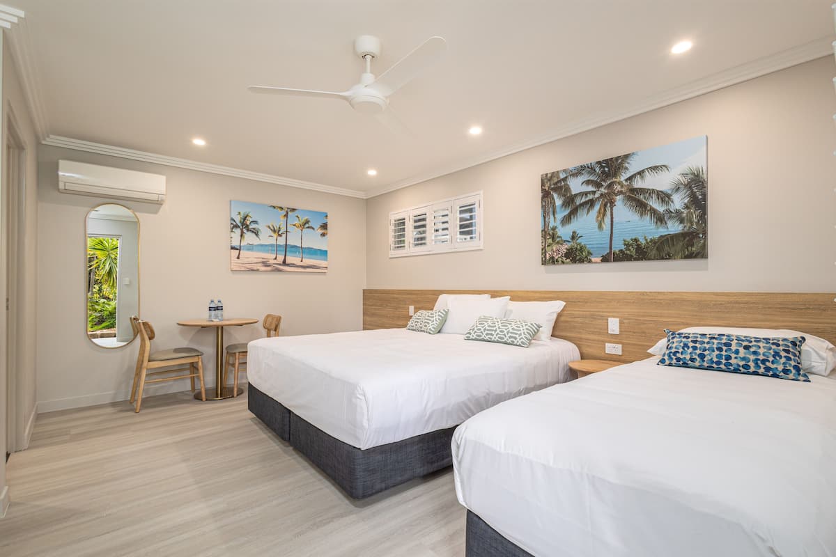 A hotel room with two beds, a small table with chairs, a ceiling fan, and artwork on the walls.