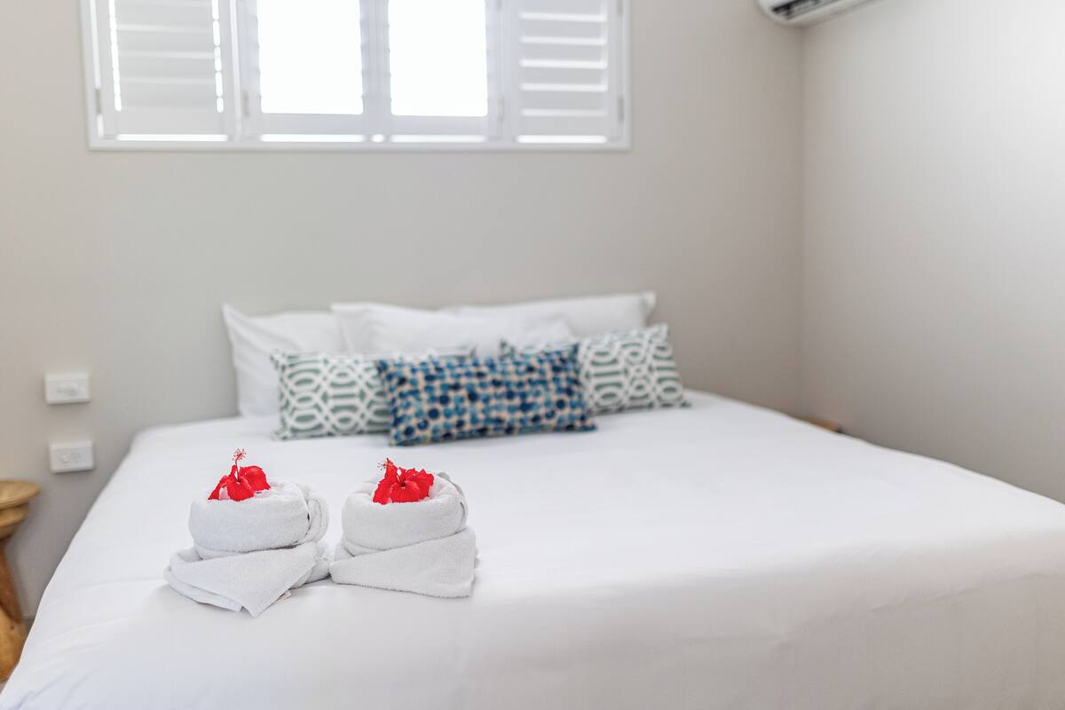 A neatly made bed with white linens and two towels with red flowers, patterned pillows, and a window with blinds.