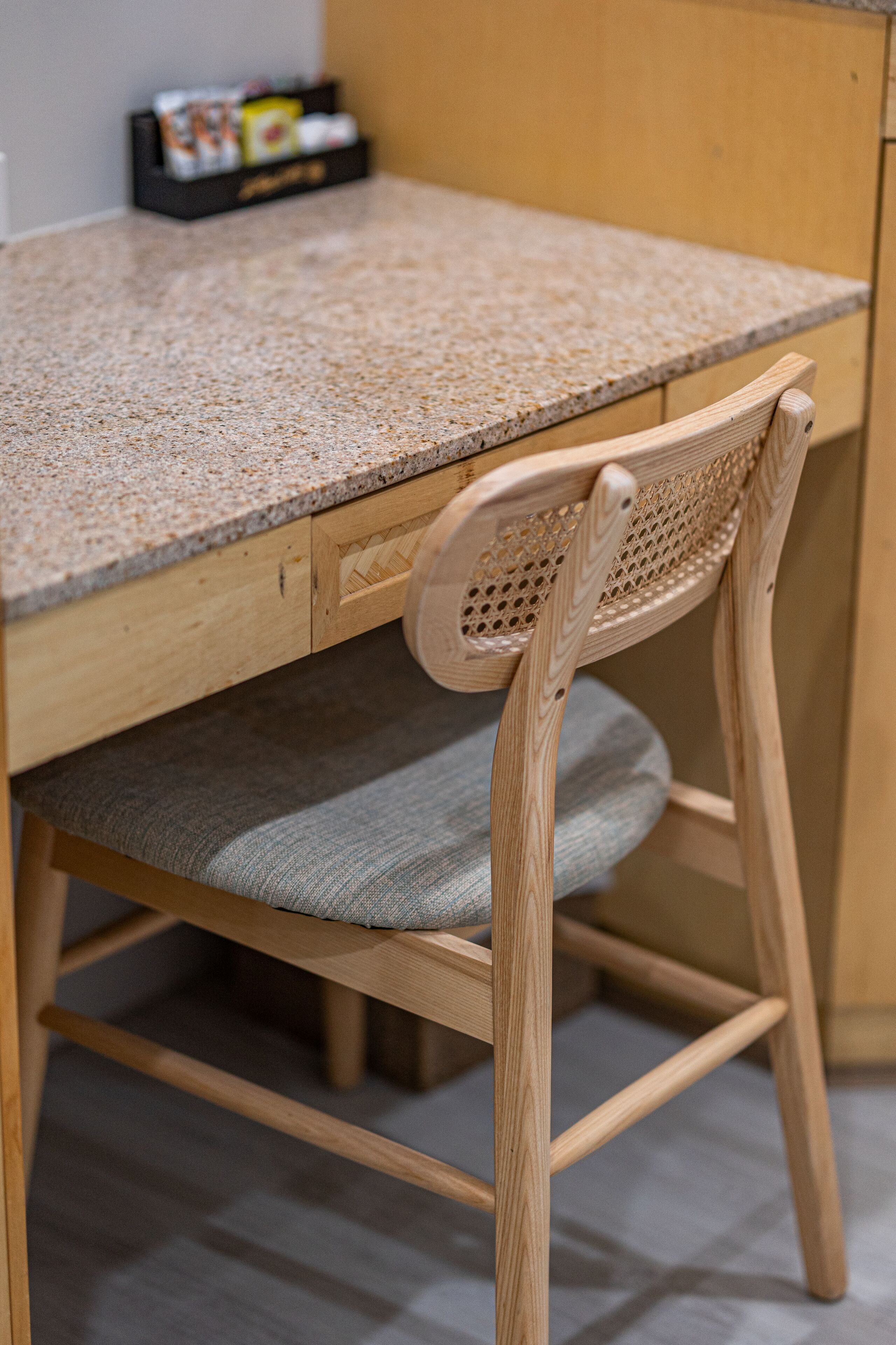 A wooden desk with a grey cushioned chair, a drawer, and a small black box on the desk.