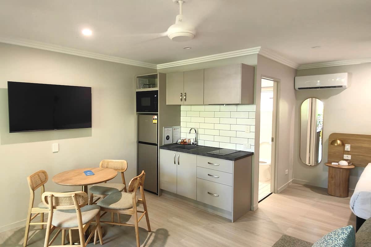 A compact hotel room with a kitchenette, a dining area with a table and chairs, a bed, and a television mounted on the wall.