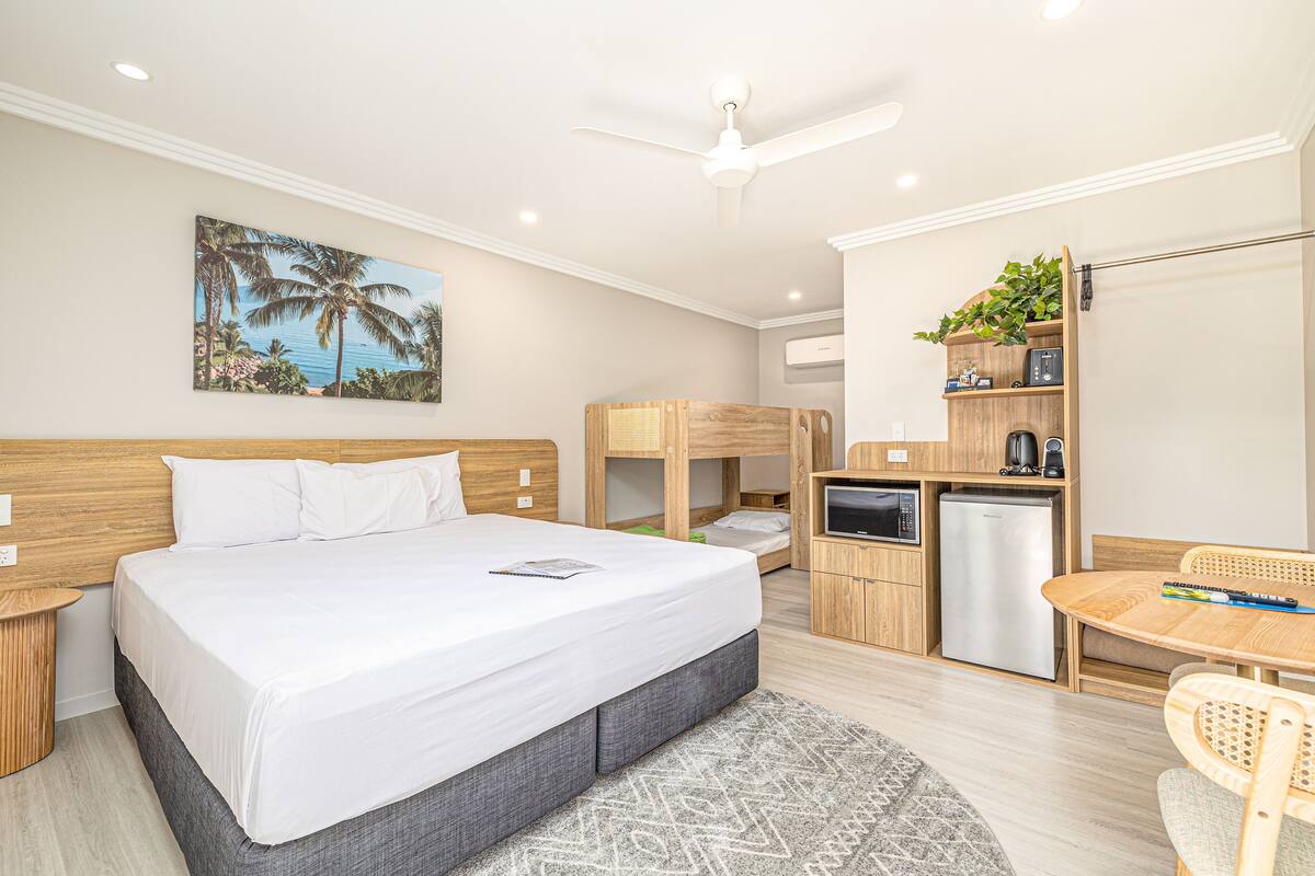 A bedroom with a large bed, a desk with a chair, a small kitchenette, and a ceiling fan.